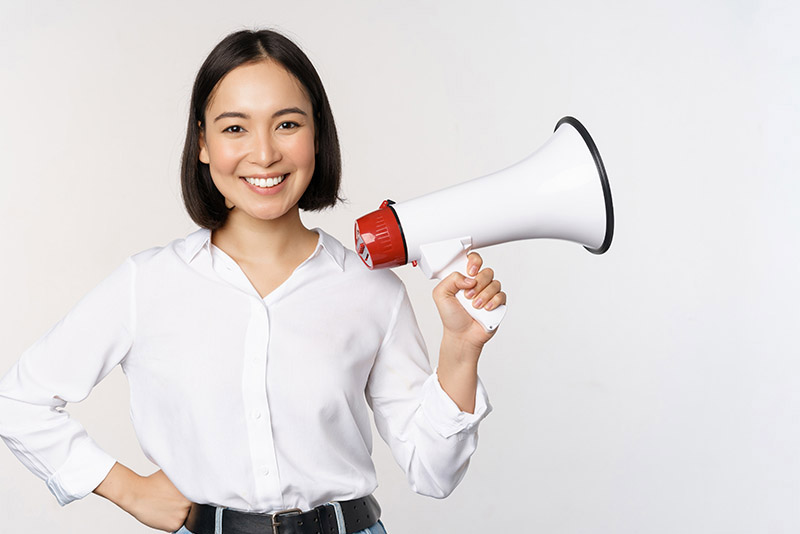 Smiling young asian woman posing with megaphone, concept of news, announcement and information, standing over white background.